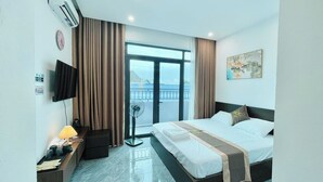 A hotel room with a bed, a TV, a desk with a lamp, a fan, and a view of the outside. - Moc Hotel Hung Yen (Nguyen Van Linh)