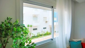 A room with a window overlooking a building with multiple windows and balconies. There is a potted plant in the foreground and a cushion on the right side of the window.