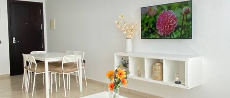 A modern living room with a white sofa, yellow and blue cushions, a white coffee table with a vase of flowers, a white dining set, and a chandelier.