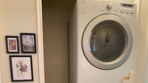 A laundry room with stacked front-loading washers and dryers, a trash can, and framed pictures on the wall. - Teal Door Rice Village Retreat World Cup dates open (Houston)