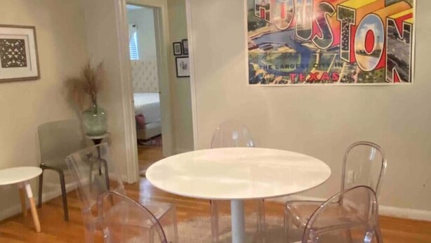 A dining area with a round table and four clear chairs, a ceiling light fixture, and a 'Houston' poster on the wall. - Teal Door Rice Village Retreat World Cup dates open (Houston)