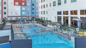 Une piscine entourée d’une clôture, avec des chaises longues et un espace dédié au bronzage, le tout bordé par un bâtiment doté de nombreuses fenêtres et de balcons.