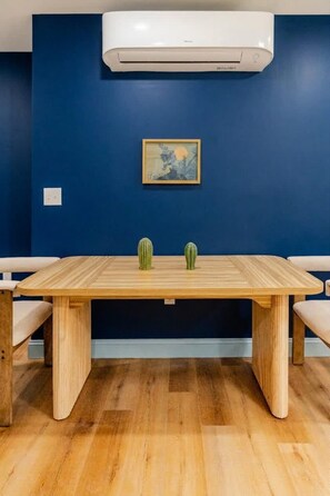 A room with a wooden table, two green cacti, a painting on the wall, and an air conditioning unit above.