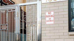 A building with a white brick facade and a metal gate with a 'NO PARKING ON SIDEWALK' sign.