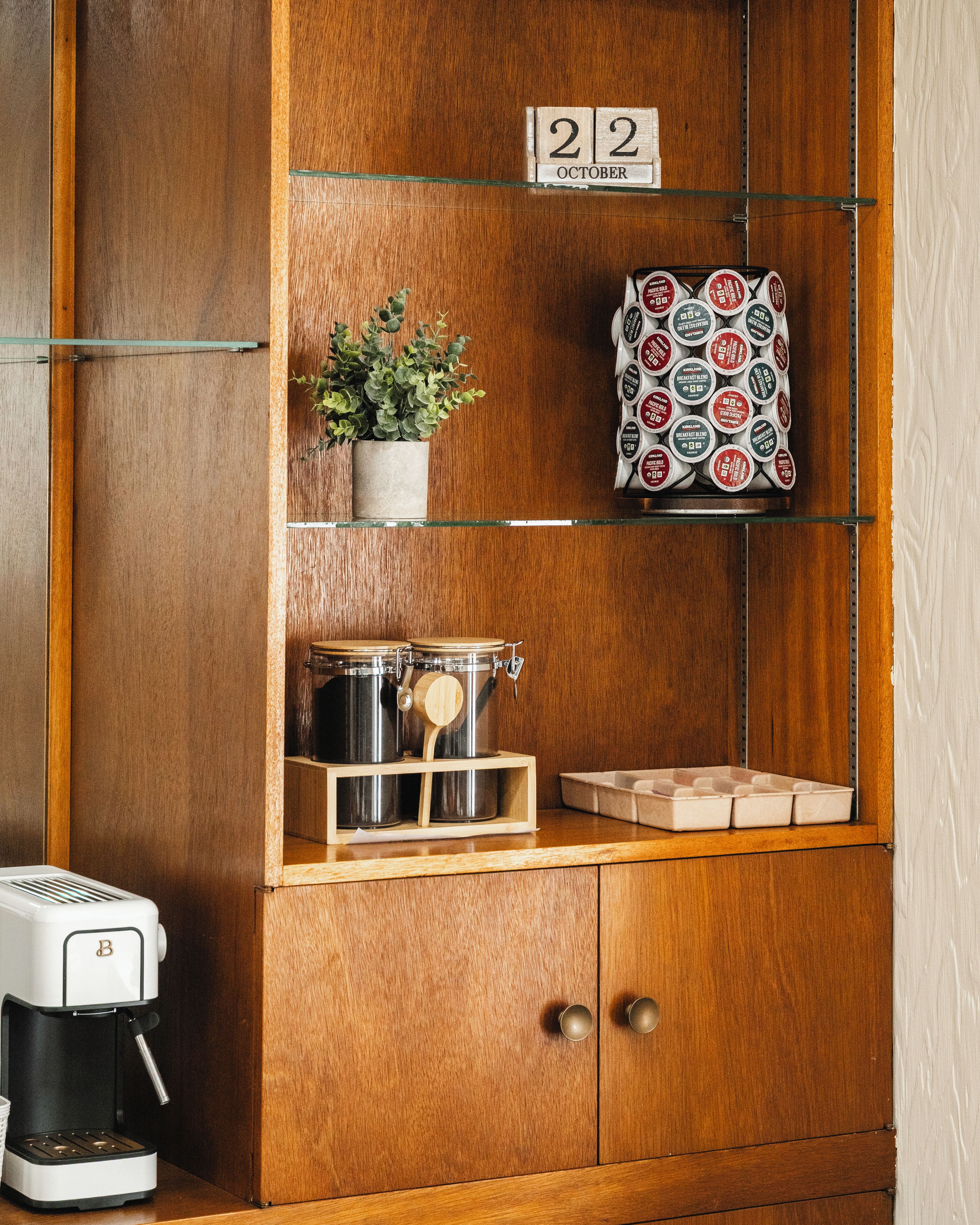 Un mueble de madera con estantes que contiene una planta en maceta, cápsulas de café, una cafetera y un cartel con la fecha.
