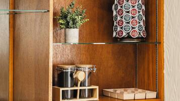Un mueble de madera con estantes que contiene una planta en maceta, cápsulas de café, una cafetera y un cartel con la fecha.