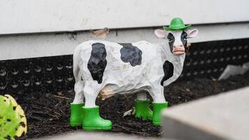 Una estàtua decorativa de vaca portant un barret verd i botes, col·locada sobre una superfície amb un ocell a prop.