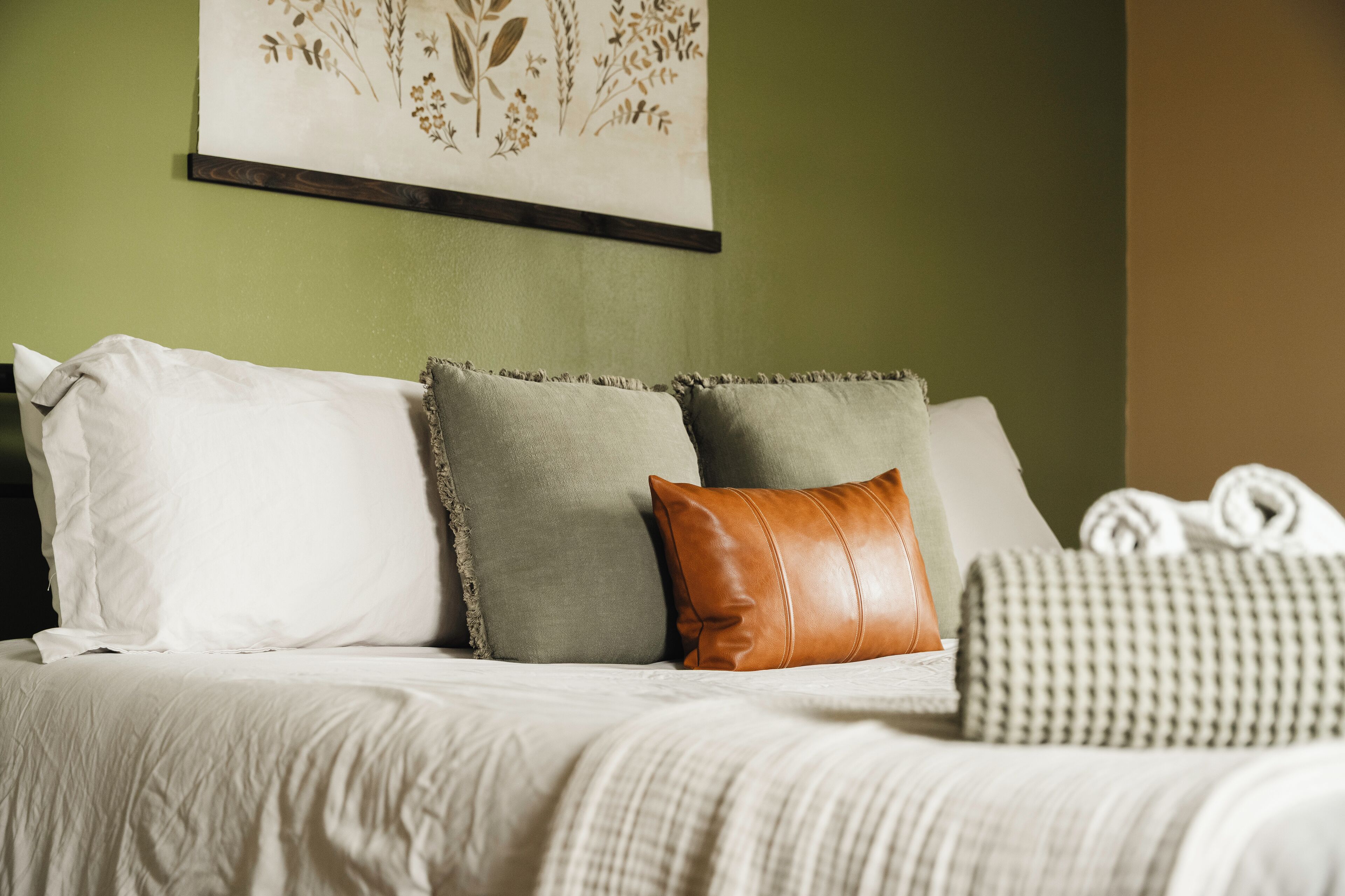 Una cama con almohadas blancas y verdes, una almohada de cuero marrón y una almohada cuadriculada.