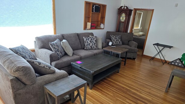 A living room with two grey sofas, a coffee table, and a wooden floor. There is a wooden shelf with books and a clock on the wall.