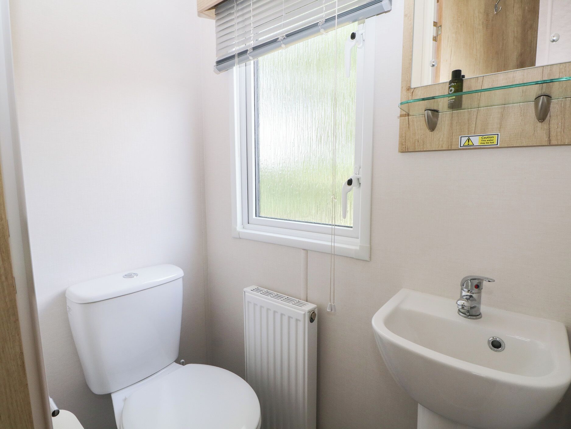 A small, clean bathroom with a toilet, a radiator, a window with blinds, and a sink with a mirror above it.