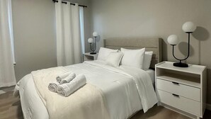 A neatly made bed with white linens and a rolled towel, a bedside table with a lamp, and a nightstand with a lamp. - Cozy 2-bedroom apartment Near Six Flags & Attractions (Springfield)