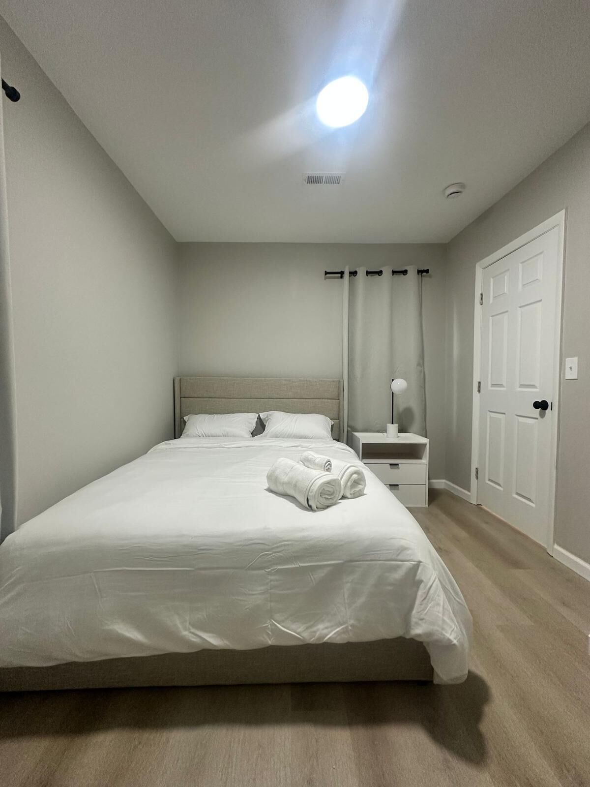 A neatly made bed with white linens and two rolled towels, a bedside table with a lamp, a door, and a ceiling light.
