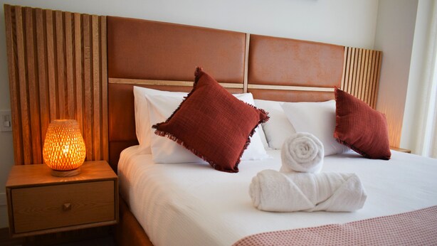 A neatly made bed with a brown headboard, two red pillows, a white pillow, and a rolled white towel. There is a wooden nightstand with a lamp beside the bed.