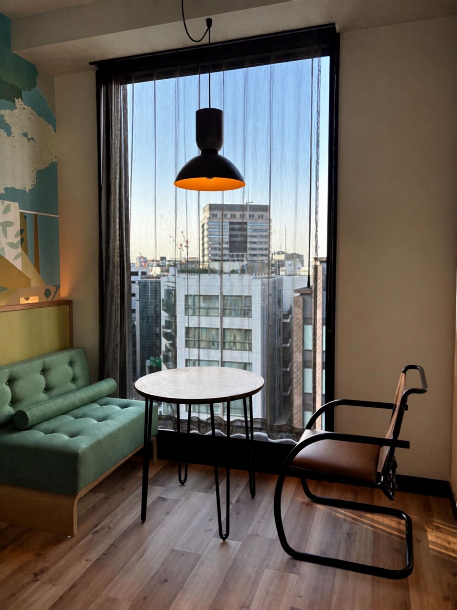 A room with a green sofa, a round table, a chair, and a large window with a view of buildings.