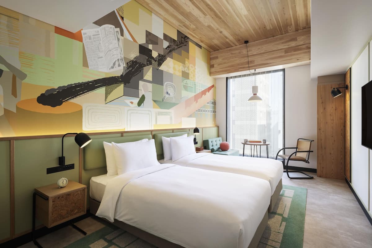 Une chambre d’hôtel avec deux lits, un plafond en bois, une grande fenêtre, un téléviseur et un mur décoré d’œuvres d’art abstraites.