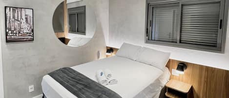 Quarto moderno com cama, mesas de cabeceira, espelho e janela com persianas.