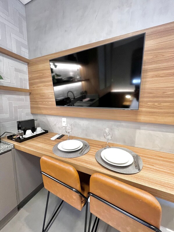A modern kitchen with a wooden countertop, two bar stools, a TV mounted above the counter, and a small coffee station. - AD 330 (São Paulo)
