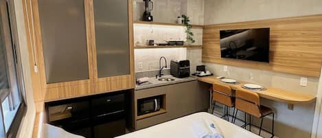 A compact hotel room with a bed, a desk, a kitchenette, and a TV mounted on the wall.