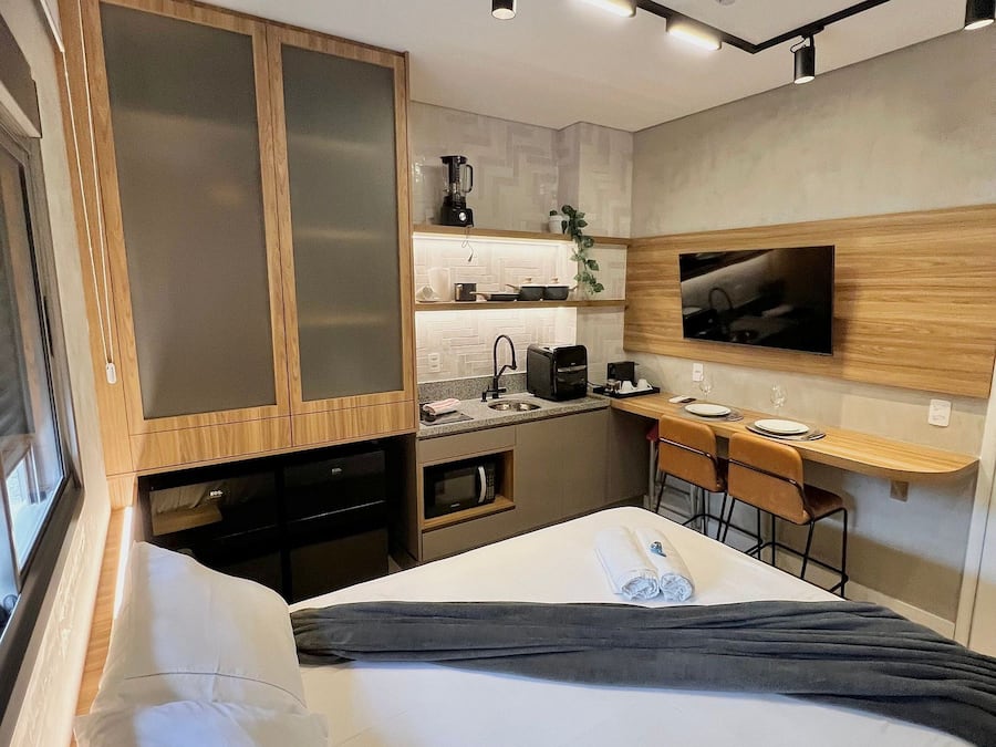 A compact hotel room with a bed, a desk, a kitchenette, and a TV mounted on the wall.