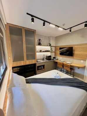 Quarto de hotel pequeno com uma cama, escrivaninha, cozinha compacta e televisão fixada na parede.