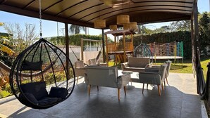 Zona d'estar a l'aire lliure coberta amb mobles moderns, una cadira penjant i una zona de jocs al fons.