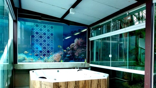Un aqüari gran amb peixos tropicals i corall, una banca de fusta i una estructura de vidre amb vistes a vegetació exterior.