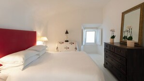 Un dormitorio con cabecera roja, cama con ropa blanca, mesita de noche con lámpara, mesa con un farol decorativo, cómoda con espejo y ventana con vistas.