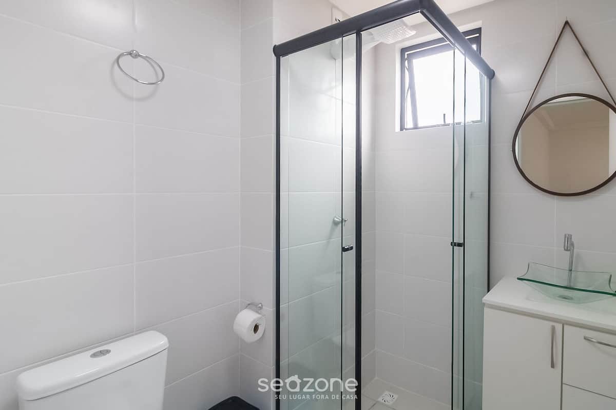 A modern bathroom with a glass shower enclosure, a toilet, a glass sink, and a round mirror on the wall.