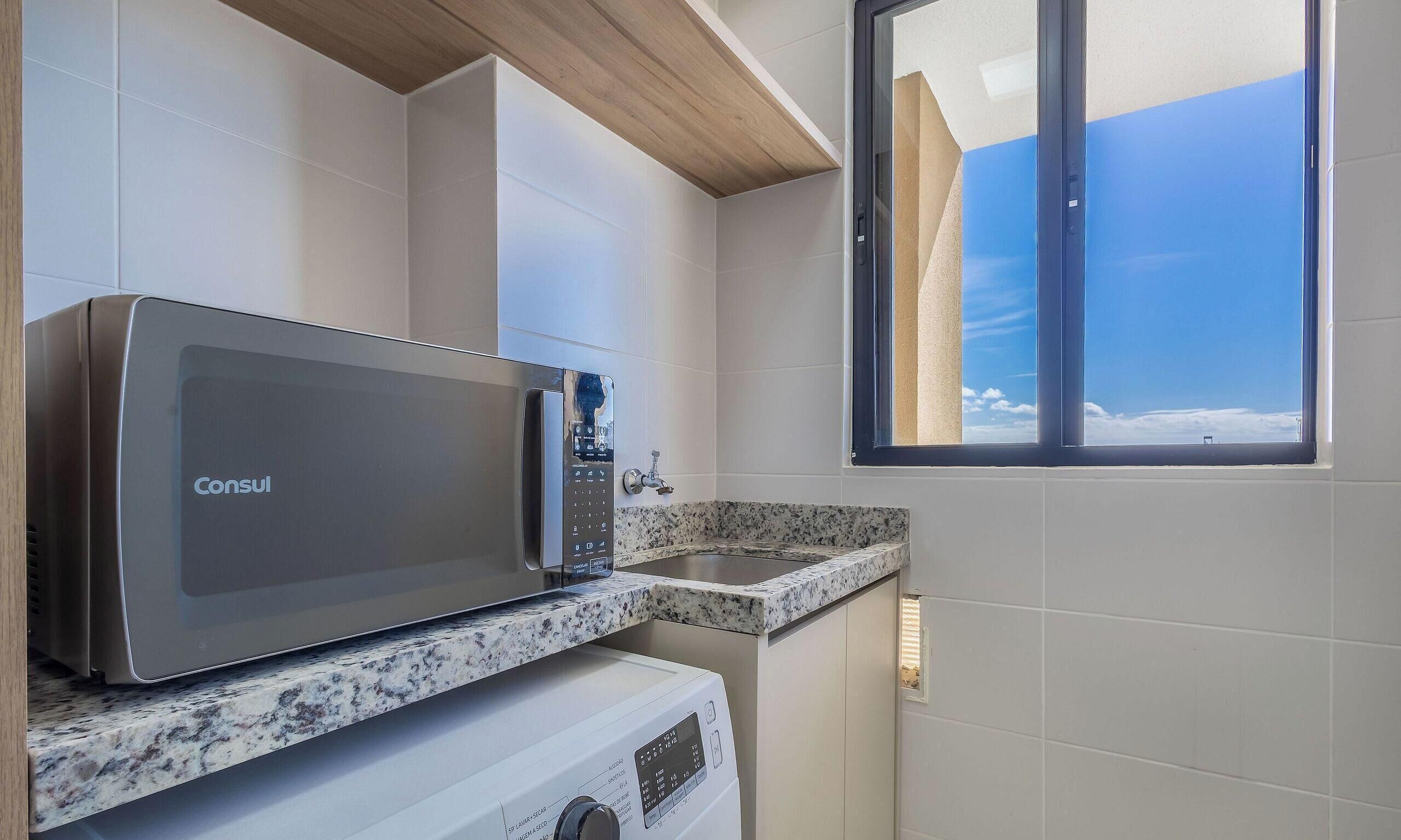 A compact kitchen with a stainless steel microwave, a built-in oven, and a window with a view of the sky.