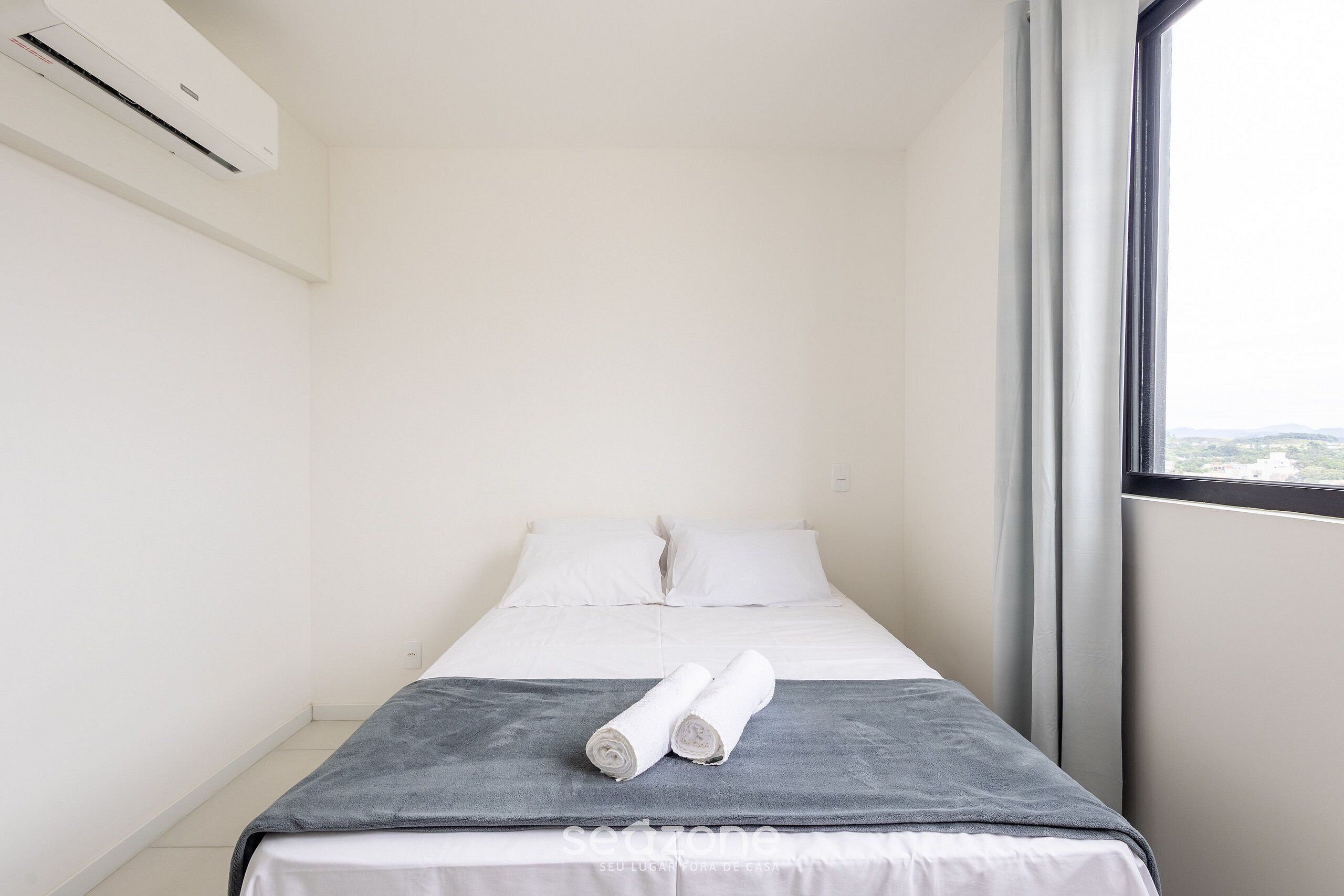 A hotel room with a bed, a grey blanket, two rolled towels, white pillows, and a window with a view.