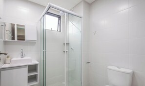 Un baño moderno con ducha envuelta en cristal, azulejos blancos, un lavamanos con espejo y un inodoro.