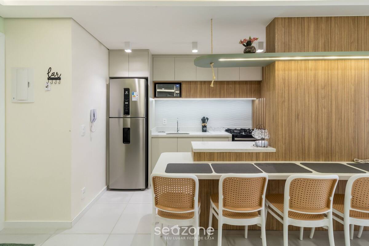 A modern kitchen with a stainless steel refrigerator, a microwave, and a wooden accent wall. There is a dining area with a white table and wicker chairs.