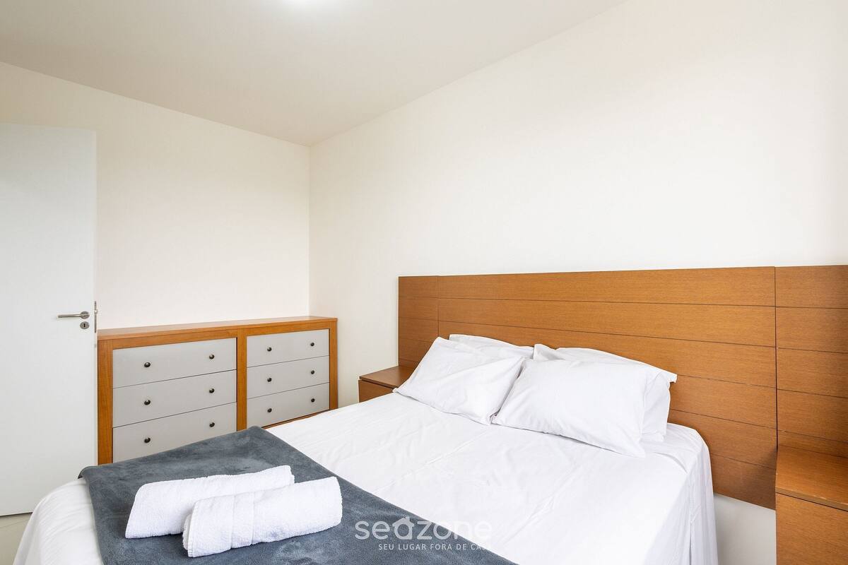 A neatly made bed with white linens and a gray throw blanket, a wooden headboard, and a nightstand with a drawer unit beside it.