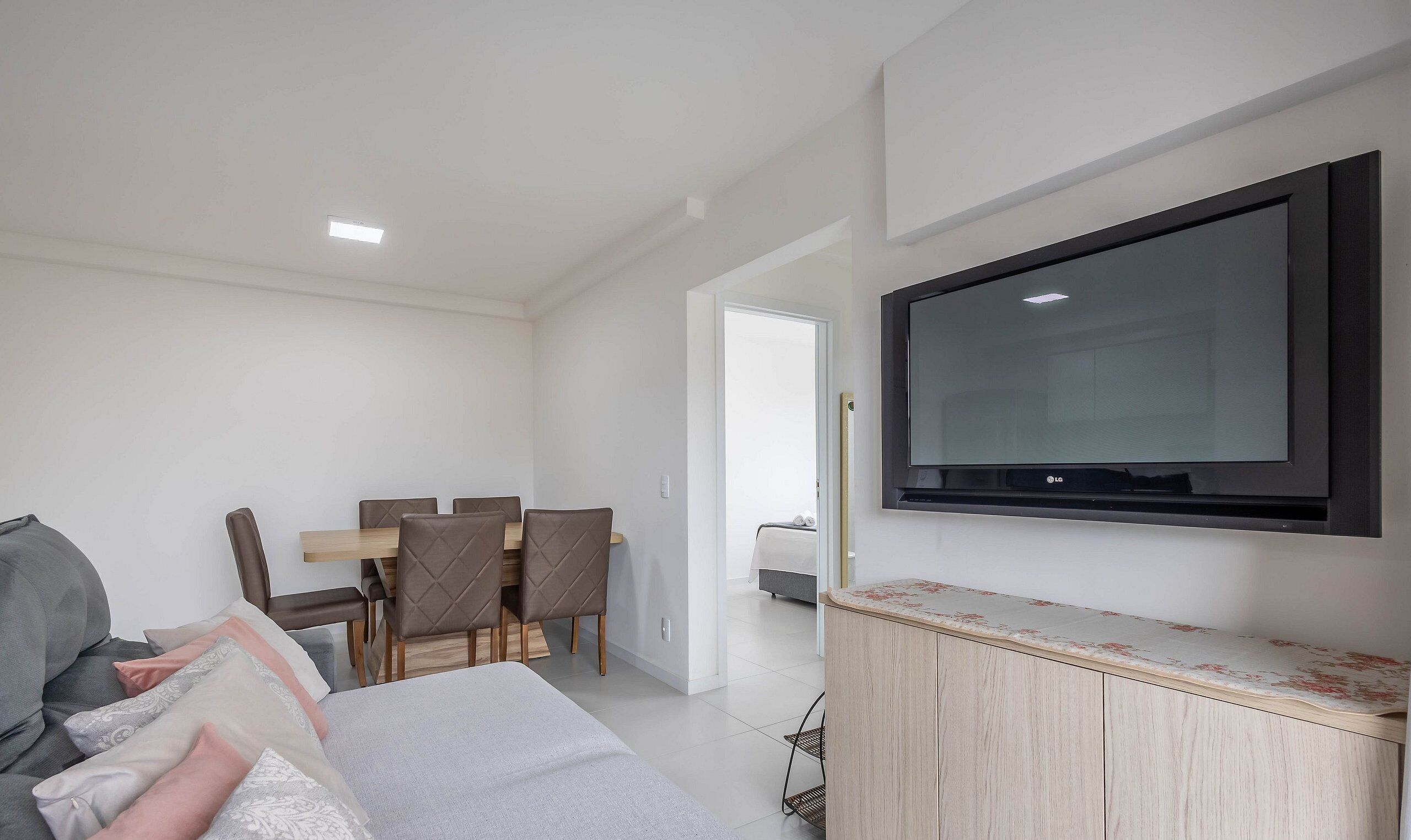 A modern, well-lit room with a flat-screen TV mounted on the wall, a dining area with a table and chairs, and a bed with a headboard.