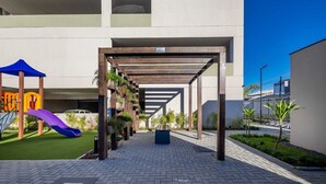 Un espace extérieur abrité doté d’une aire de jeux, comprenant un toboggan et des structures d’escalade, attenant à un bâtiment agrémenté d’une pergola et de plantes en pot.