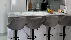 A modern kitchen with white cabinets, a marble countertop, and a built-in refrigerator. There are four bar stools with black bases and grey seats.