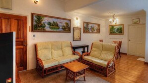 A living room with a sofa set, a coffee table, and a television. There are paintings on the walls and a door in the background. - Cozy 10-bedroom bungalow in Cameron Highlands (Tanah Rata)