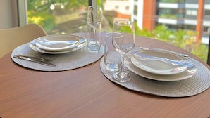 Una mesa de comedor con platos, vasos y cubiertos para dos personas, ubicada cerca de una ventana con vistas a edificios y vegetación.