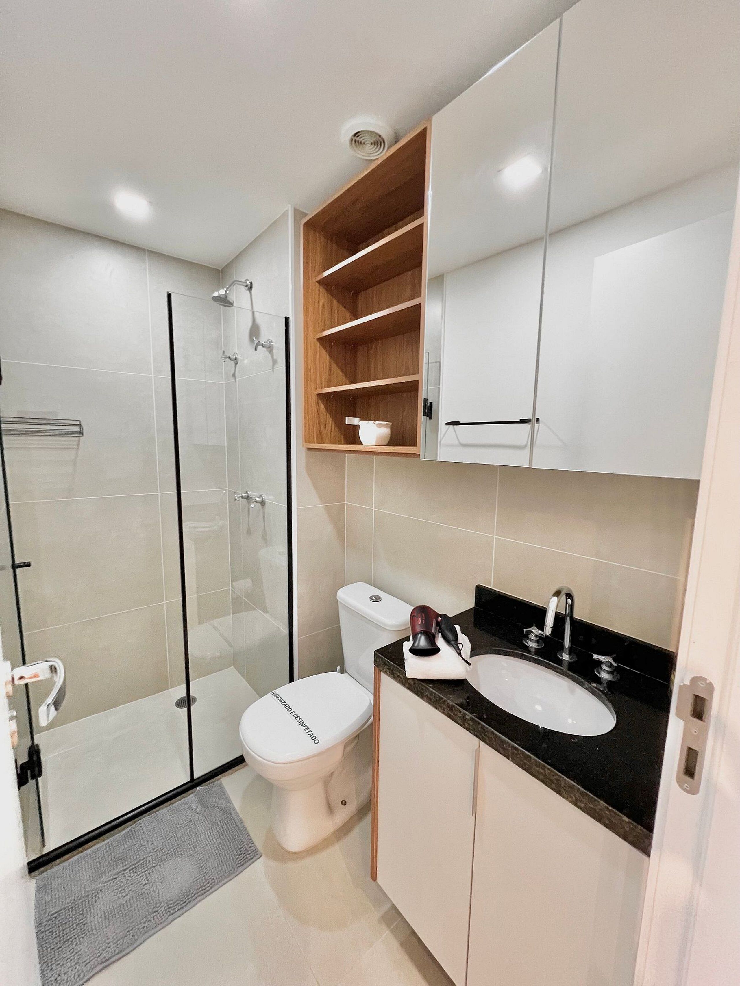 Un baño moderno con ducha de vidrio, inodoro blanco, lavamanos con encimera negra y estantes de madera sobre el lavamanos.
