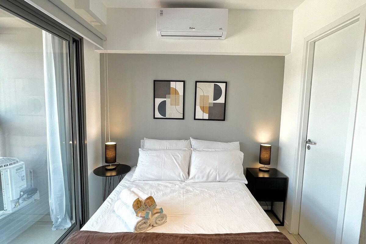 Une chambre d’hôtel comprenant un lit, deux tables de chevet, un appareil de climatisation, une fenêtre avec des rideaux et deux œuvres d’art abstraites accrochées au mur.