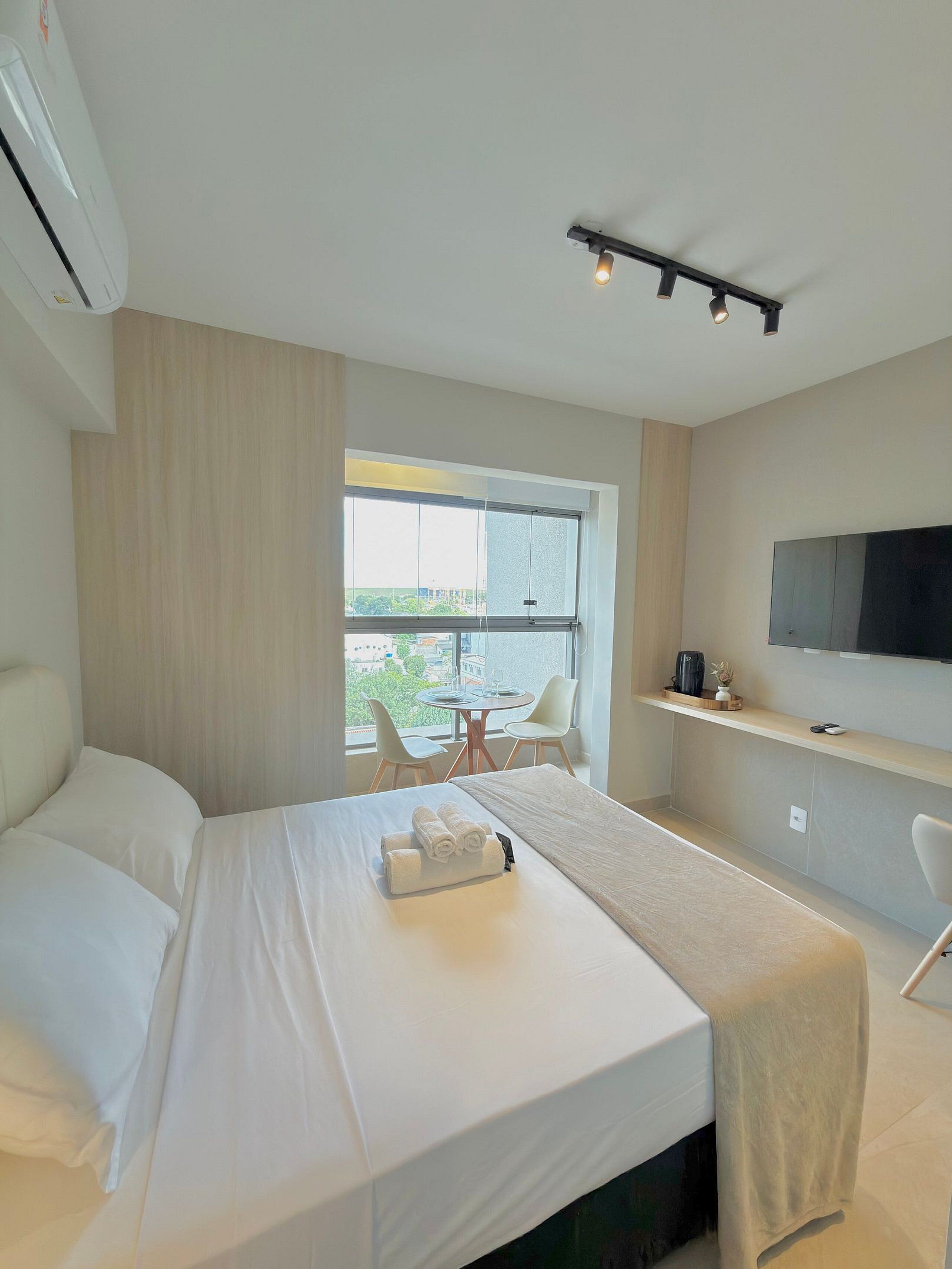 A hotel room with a bed, a TV mounted on the wall, a small table with chairs, and a window with a view of the city.