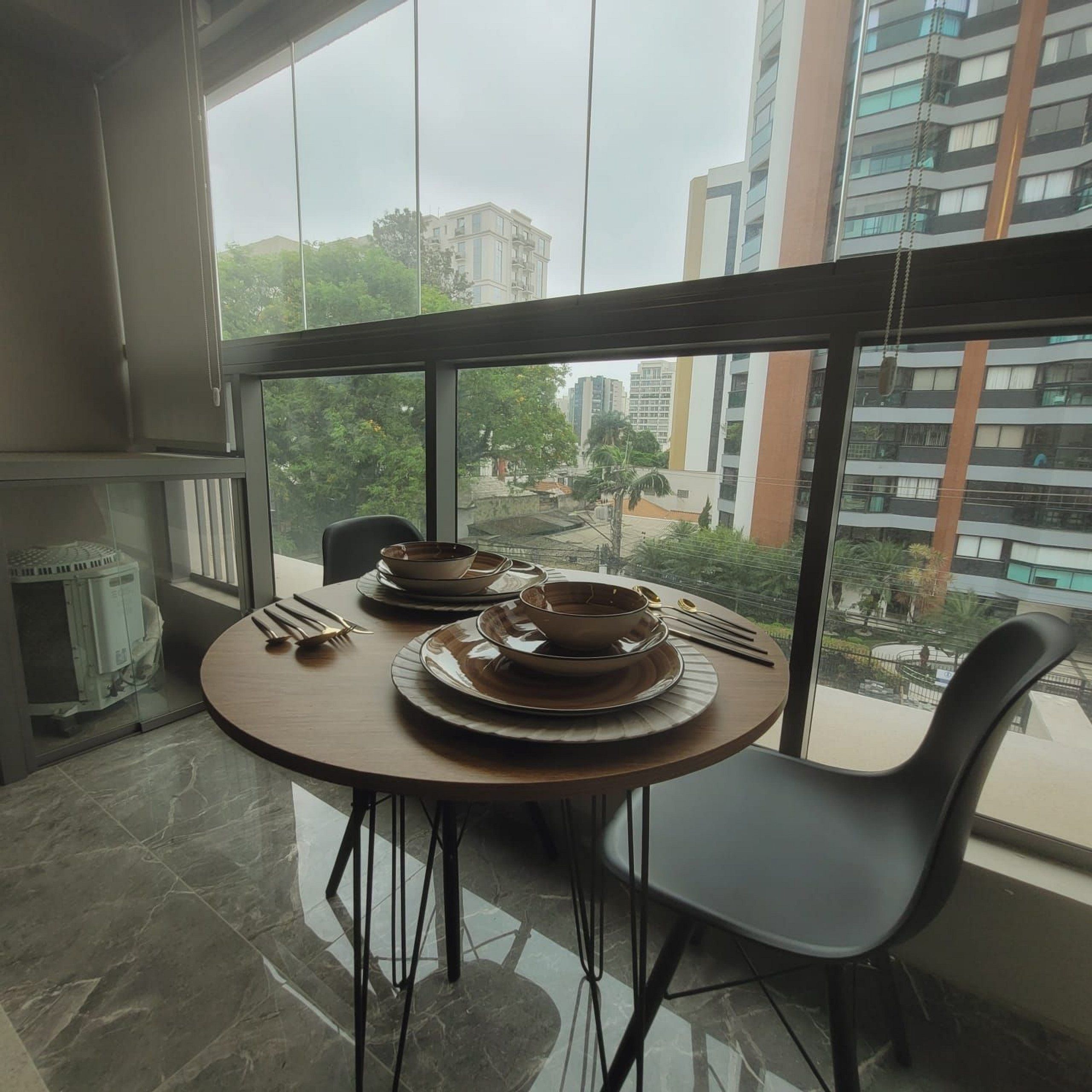 A dining area with a table set for two, a chair, and a view of buildings through large windows.
