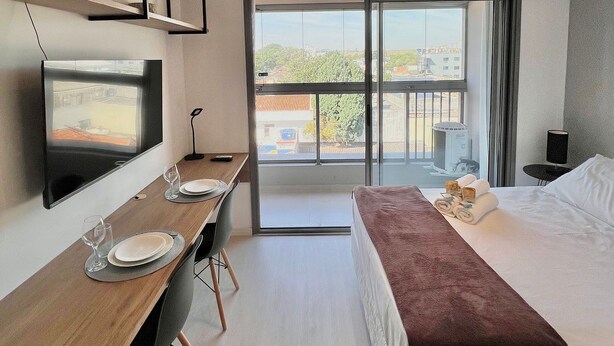 Habitación de hotel moderna con cama, escritorio con televisor, zona de comedor y balcón con vistas.