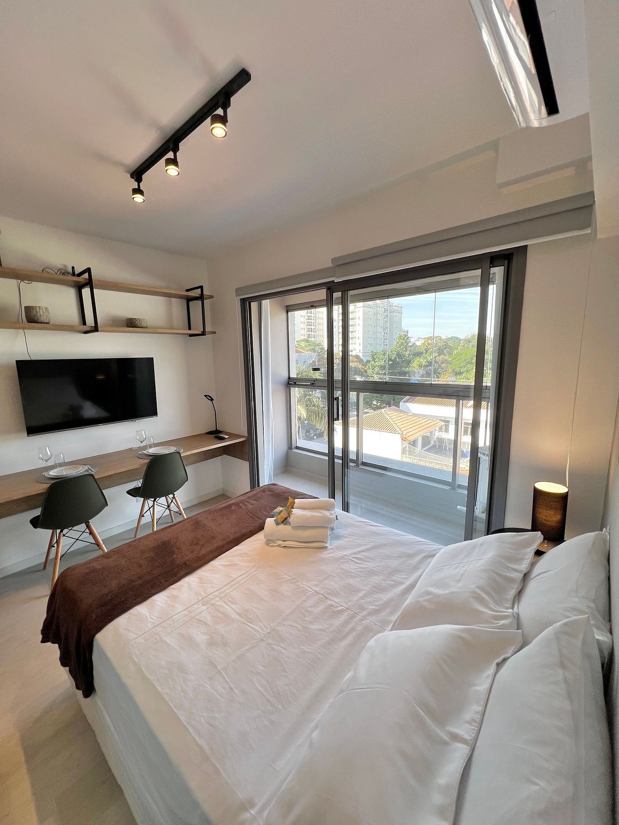 A modern hotel room with a large bed, a desk with two chairs, a flat-screen TV, and a balcony with a city view.