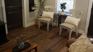 A cozy living room with a wooden floor, white chairs, a brown leather sofa, a ceiling fan, and a small side table with a plant. - Cozy Greenville Cottage. Close to downtown! Pet friendly! (Greenville)