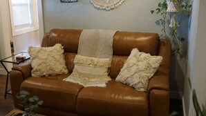 A brown leather sofa with three pillows and a throw blanket, a wooden coffee table, a macrame wall hanging, and two wall-mounted planters with green plants. - Cozy Greenville Cottage. Close to downtown! Pet friendly! (Greenville)