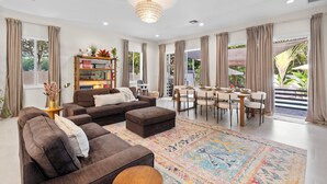 A spacious living room with a brown sectional sofa, a coffee table, and a patterned rug. There is a dining area with a table and chairs, and a bookshelf filled with books and decorative items. - New + Lux Close to everything! Gym and Pool 5 Bedrooms! (fort lauderdale)