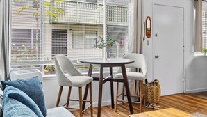 Une pièce avec une table ronde, deux chaises, une fenêtre avec des stores, une porte et un petit panier posé sur le sol.