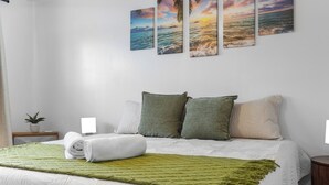 Un lit bien fait, avec une couverture verte, des oreillers blancs et une couette blanche. Au mur, on peut voir trois tableaux encadrés représentant un coucher de soleil sur l’océan.
