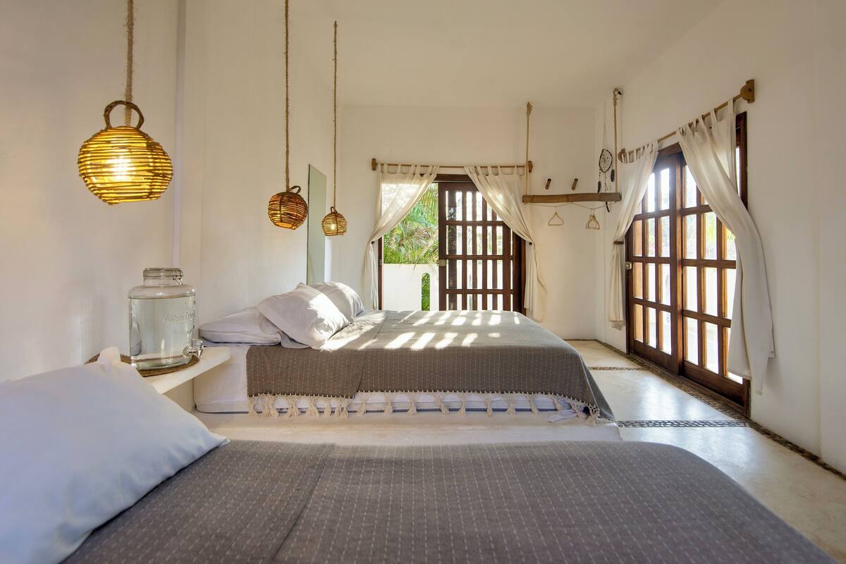 A bedroom with two beds, a hanging light, a window with curtains, and a view of greenery outside.